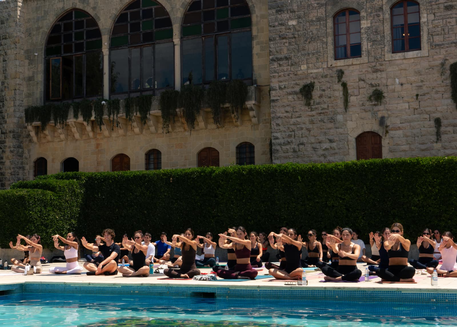 A large group seated on mats beside a pool at a previous Noonflows gathering, arms crossed in front of them mid-stretch, a stone building and tall hedge behind